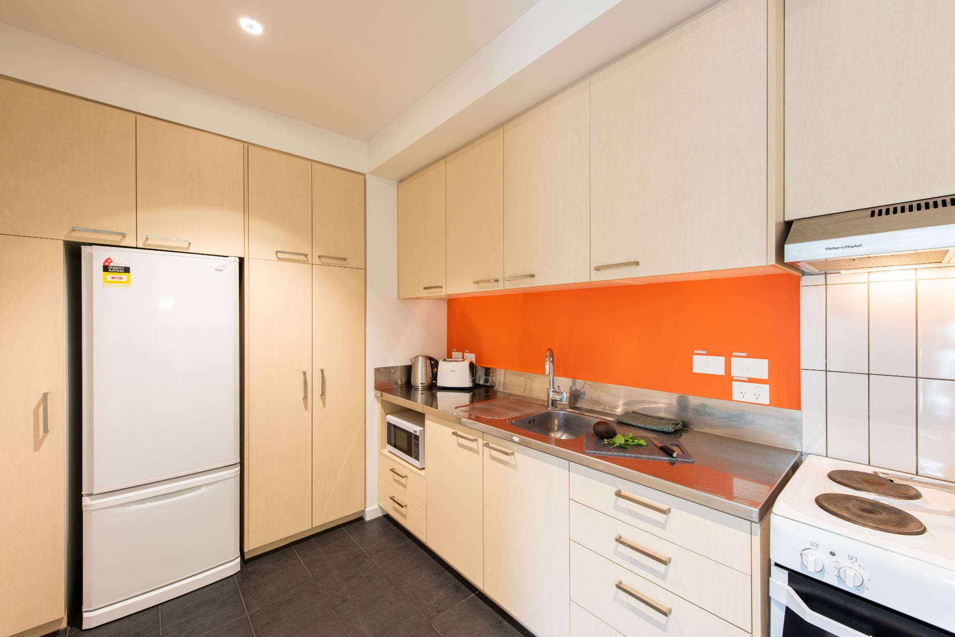 Kitchen in flat at Carlaw Park. Features light wood cabinets, fridge, stove, orange backsplash and white tile backsplash.